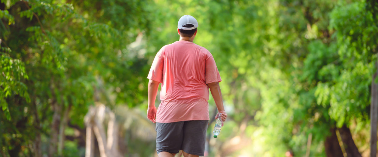 Pessoa com obesidade a caminhar num caminho rodeado de árvores verdes, vestindo uma t-shirt rosa, calções cinzentos e um boné branco, segurando uma garrafa de água.